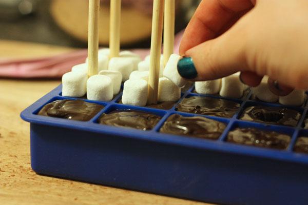 Once all chocolate has been piped, pound the tray lightly on the counter to eliminate air bubbles and to flatten the tops of the cubes if they are uneven. Working one cube at a time, insert a stick and place four mini marshmallows onto the top of the cube. 