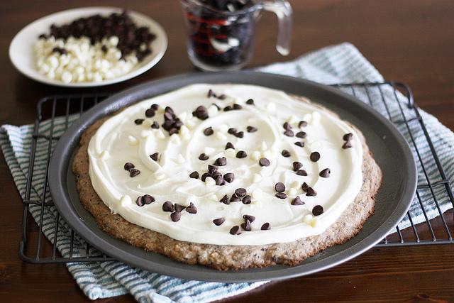 Spread mixture evenly on top of cooled cookie pizza. Sprinkle with chocolate chips and crushed cookie pieces. Melt a handful of white chocolate chips and drizzle over cookie pizza.