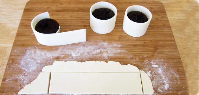 Dust work surface with cornstarch or powdered sugar. Knead and then roll out fondant to about 1/8th of an inch thickness. Cut into 2 inch by 8 1/4 inch strips, re-rolling as needed. Wrap a strip around each cupcake. Cut off any excess fondant. Brush the edges where the fondant meets with water.